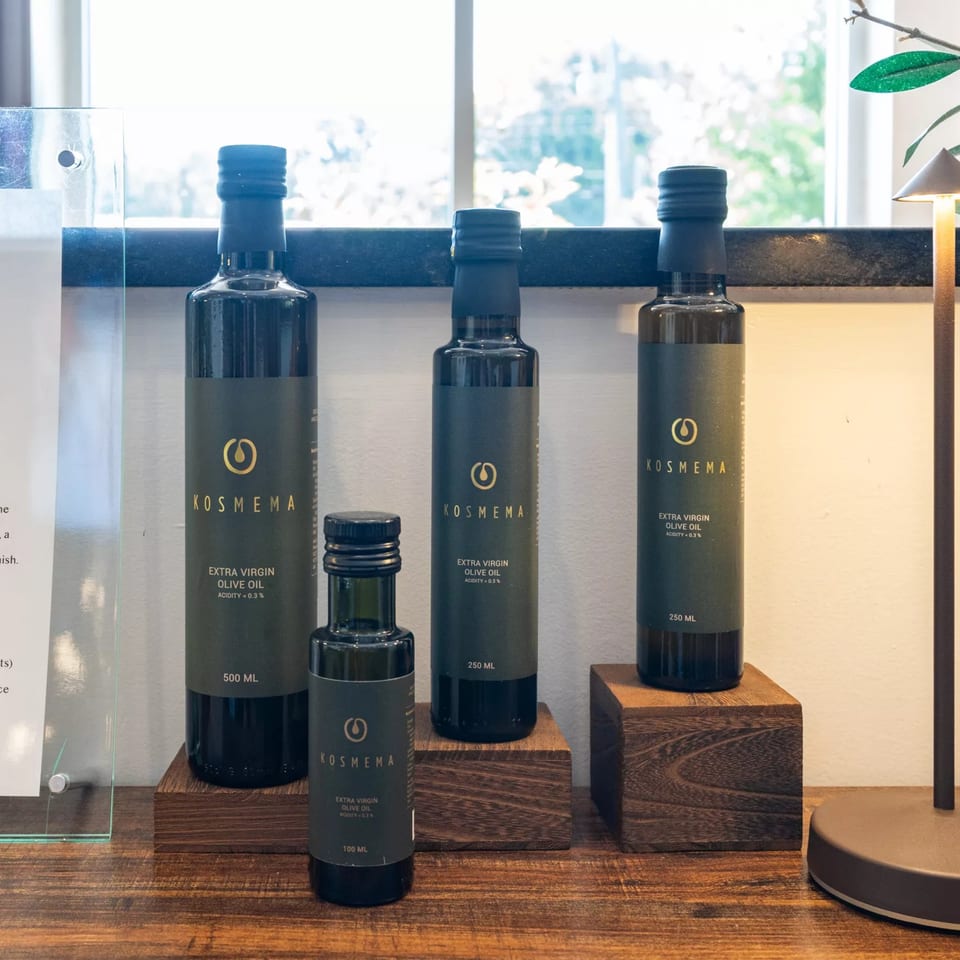Four dark bottles of Kosmema Extra Virgin Olive Oil in various sizes on wooden blocks.