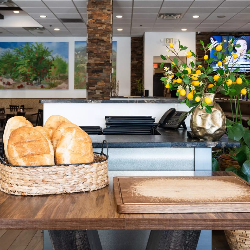 Freshly baked bread in a woven basket on a wooden table, with a restaurant counter and decor in the background.
