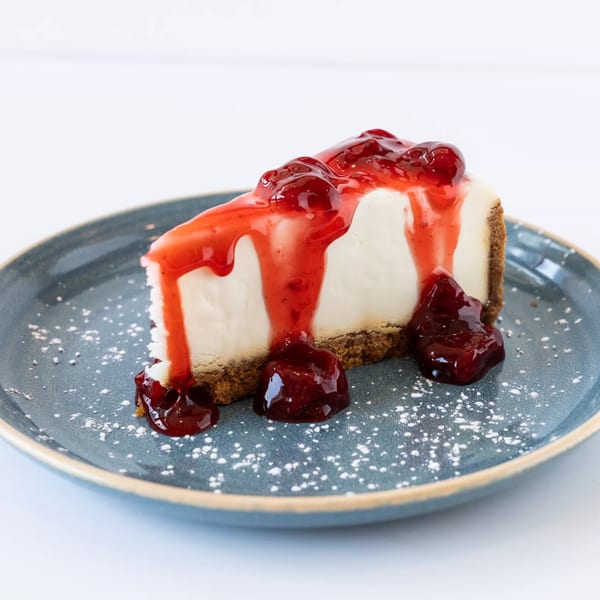 A slice of cheesecake with a graham cracker crust, topped with cherry sauce and powdered sugar.