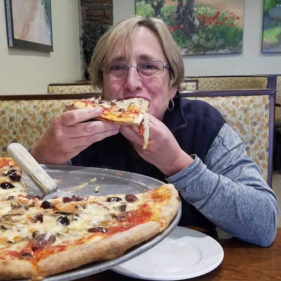 A woman in glasses happily takes a bite of a slice of pizza from a large pie.